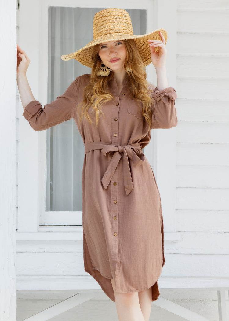 Woman wearing a brown dress and straw hat standing in front of a white wooden door.