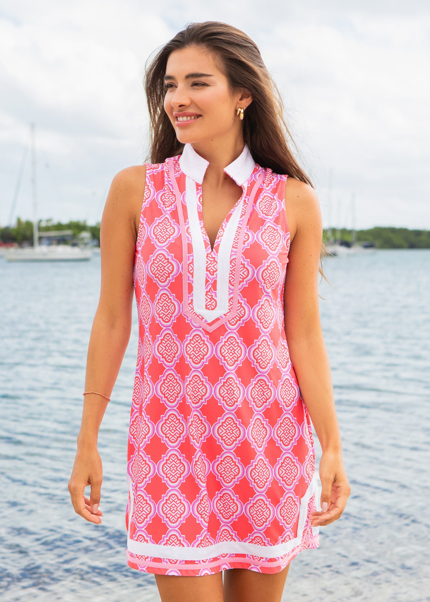 Woman wearing a pink patterned dress standing by a body of water with sailboats in the background.