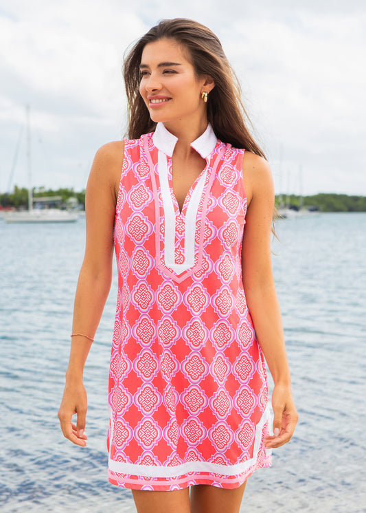 Woman wearing a pink patterned dress standing by a body of water with sailboats in the background.