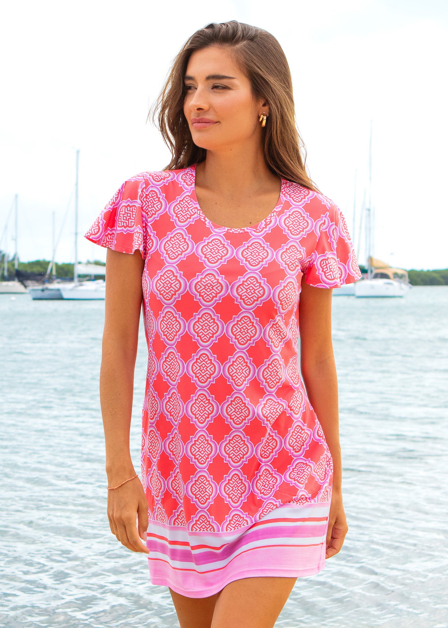 Woman wearing a pink patterned dress standing by a waterfront with boats in the background.