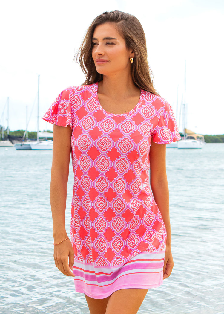 Woman wearing a pink patterned dress standing by a waterfront with boats in the background.
