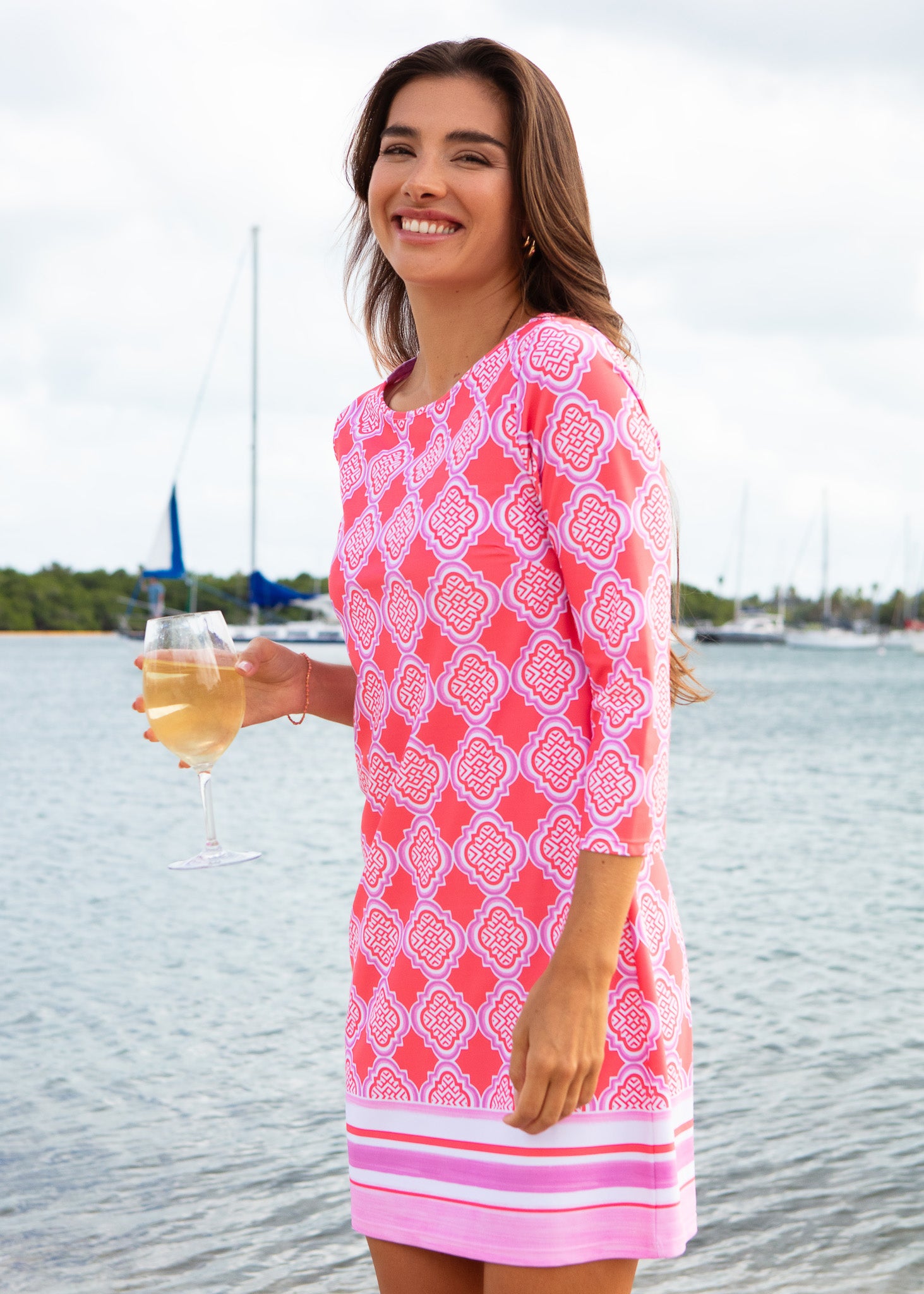 Woman in a pink patterned dress standing by a waterfront with a glass of wine.