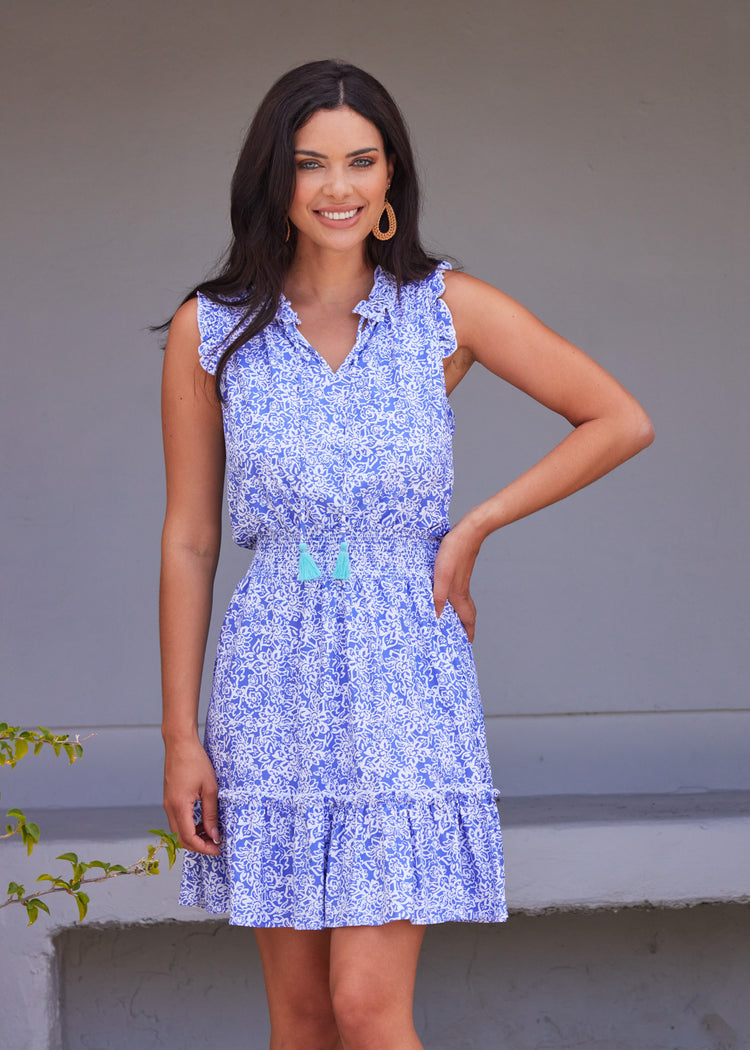 Dark haired woman wearing Lake Como Sleeveless Ruffle Dress with brown earrings and white background. 