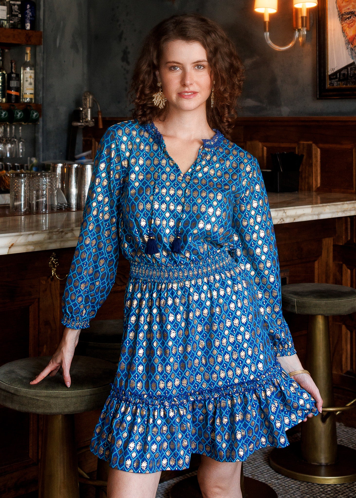 A woman wearing a blue long-sleeve dress with a gold geometric pattern, posing in front of a rustic wooden bar.