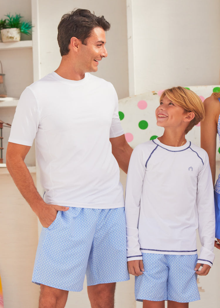 A boy and man standing in front of a wall with polka dots, wearing white long-sleeve rashguard and blue swim trunk with a geometric pattern.