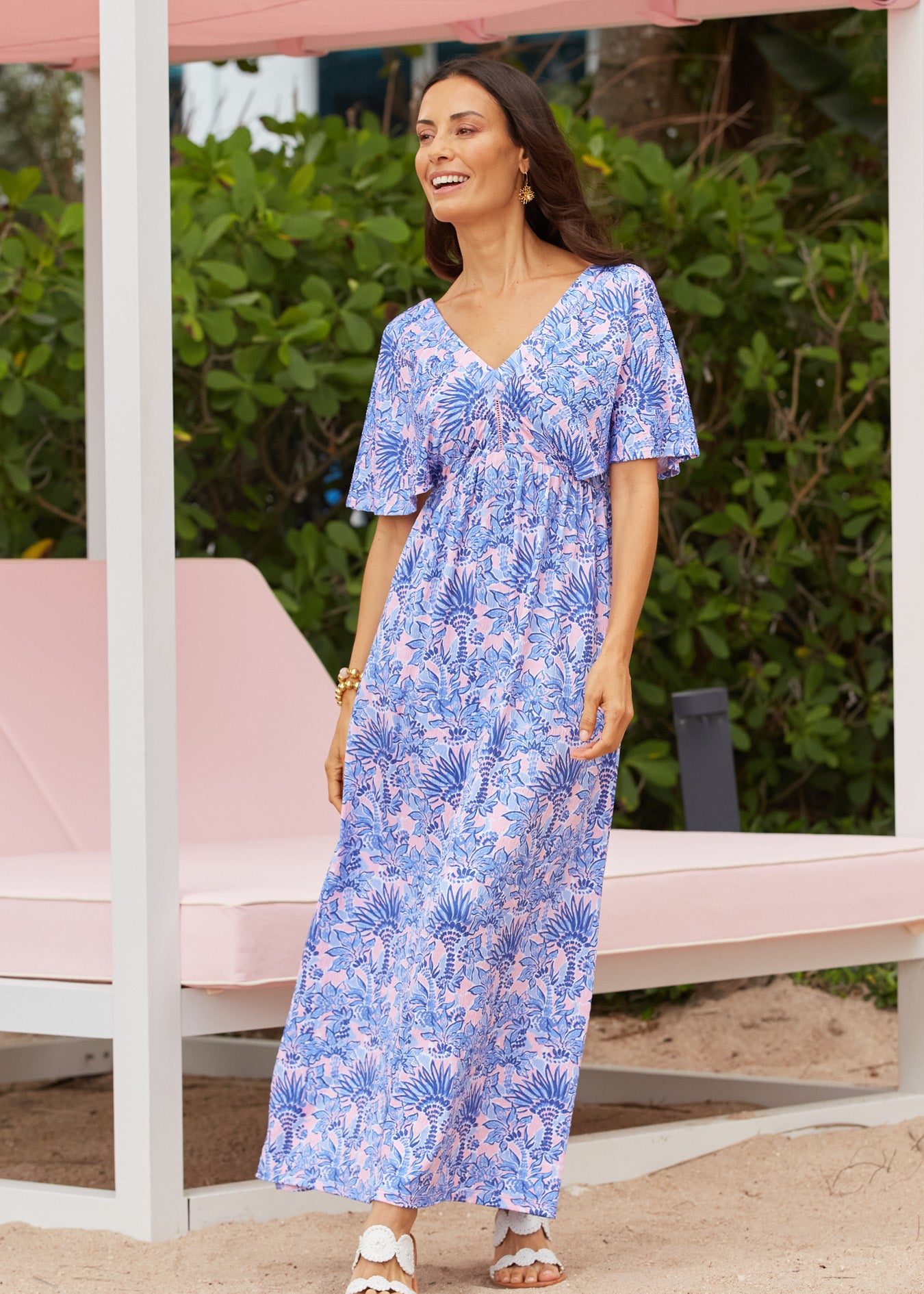 A brunette woman in front of a pink resort lounger wearing the Cabana Life sun protective & flowy Palm Beach Flutter Sleeve Maxi Dress.