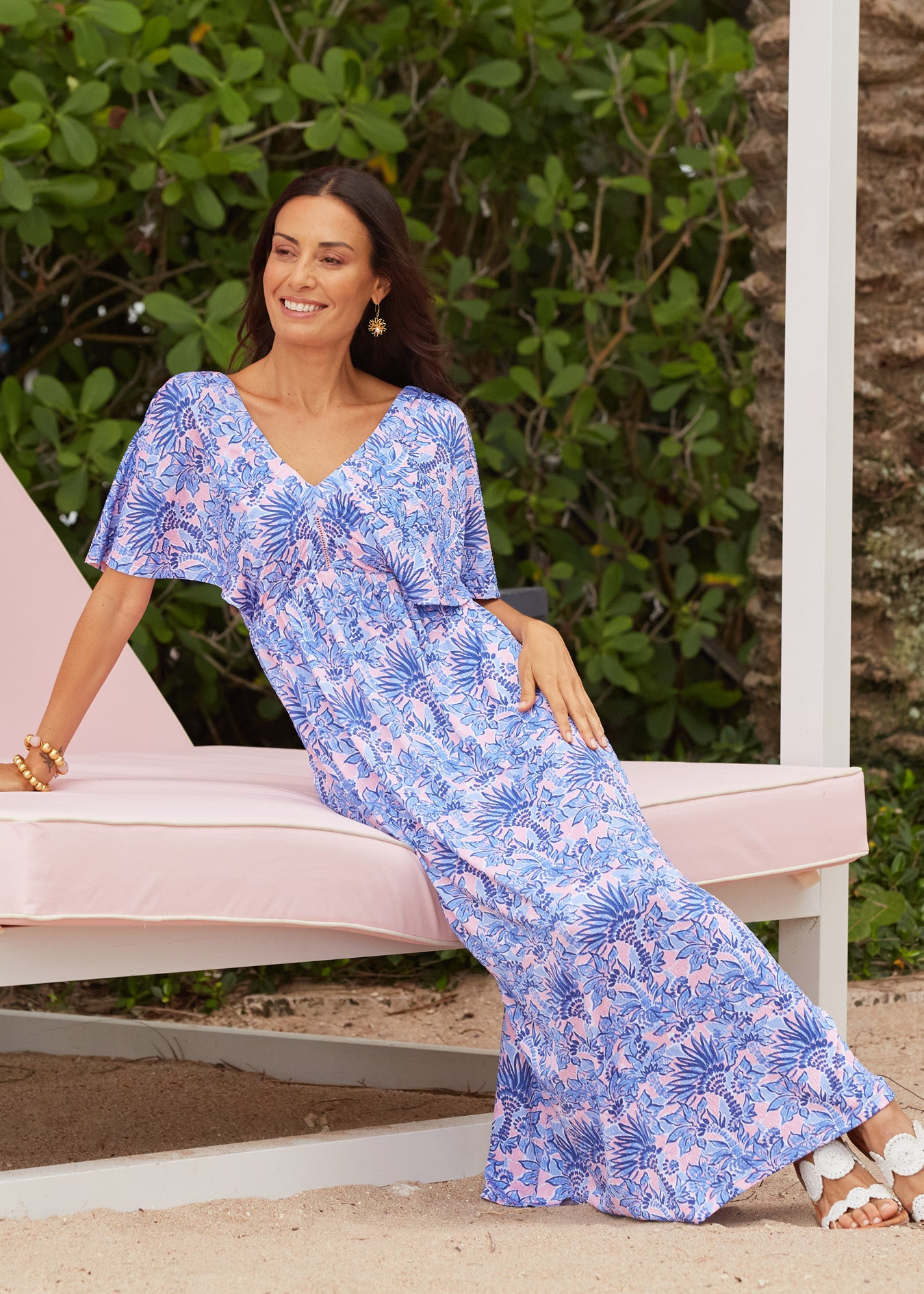 A brunette woman sitting on a pink resort lounger wearing the Cabana Life sun protective & flowy Palm Beach Flutter Sleeve Maxi Dress.