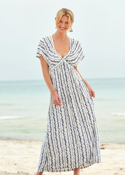 A blonde woman smiling and standing on the beach wearing the San Sebastian Flutter Sleeve Maxi Dress and navy earrings.