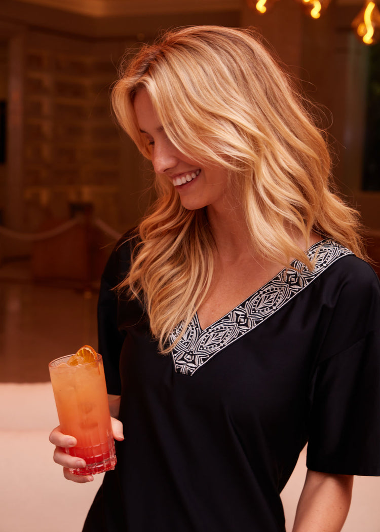 Woman looking down wearing Black Flutter Sleeve Shift Dress holding orange tropical drink.