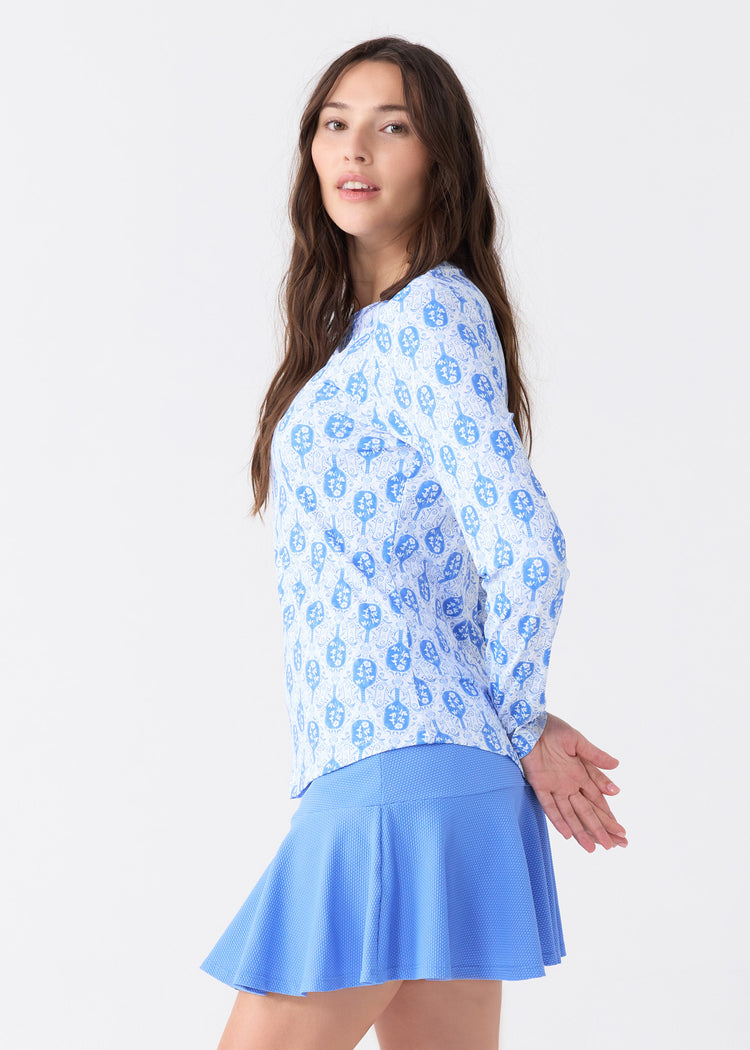 The side of a brunette woman wearing the sun safe Pickleball Long Sleeve Performance Top and Periwinkle 15" Flounce Skort from Cabana Life.