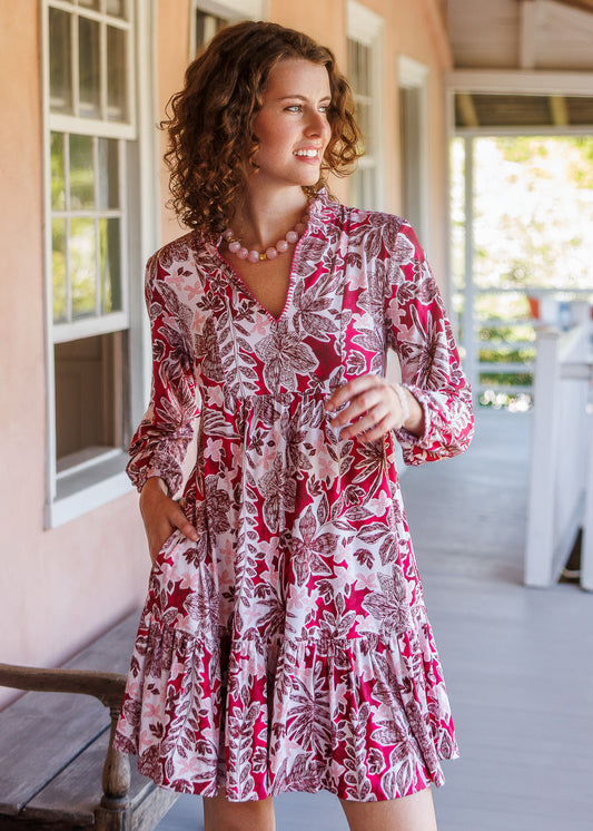 A woman wearing a pink and white tropical floral print mini dress with long sleeves and a V-neckline, standing on a porch.