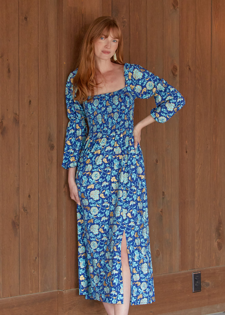 A redhead woman wearing the sun protective Navy Floral Metallic Puff Sleeve Maxi Dress from Cabana Life in front of a wood background.