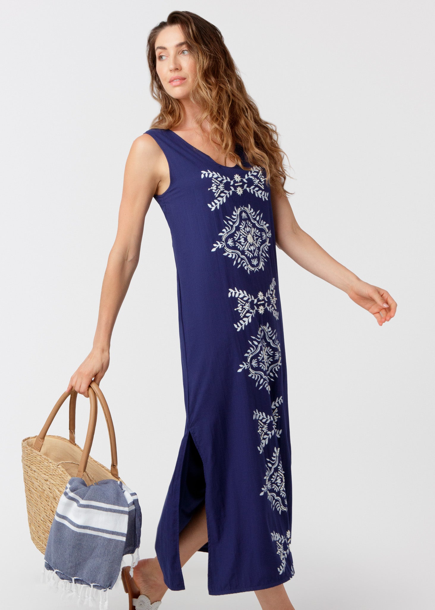 Woman wearing a navy maxi dress holding a straw bag against a white background.