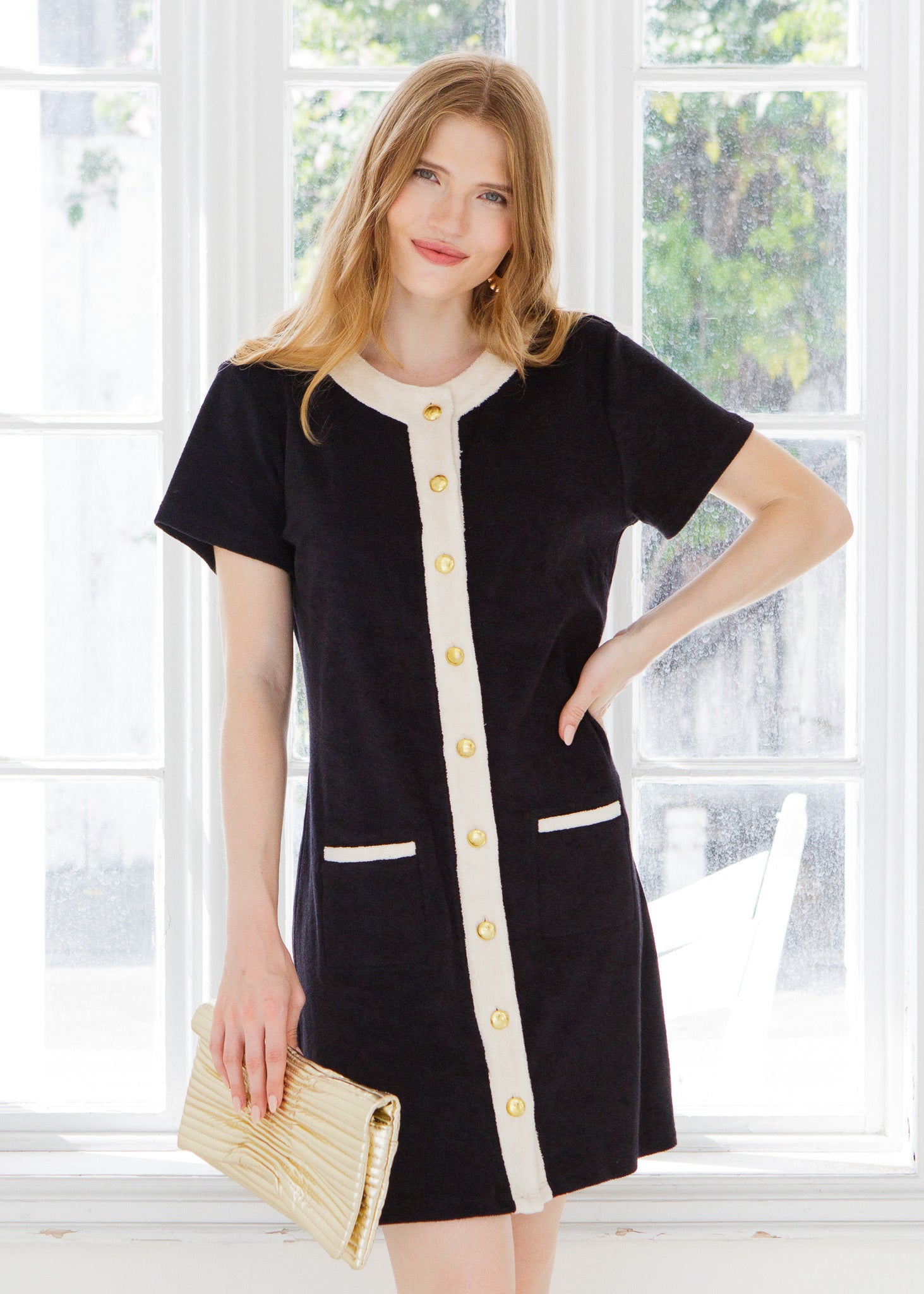 Woman wearing a black and cream terry dress with gold buttons standing outside a white window.