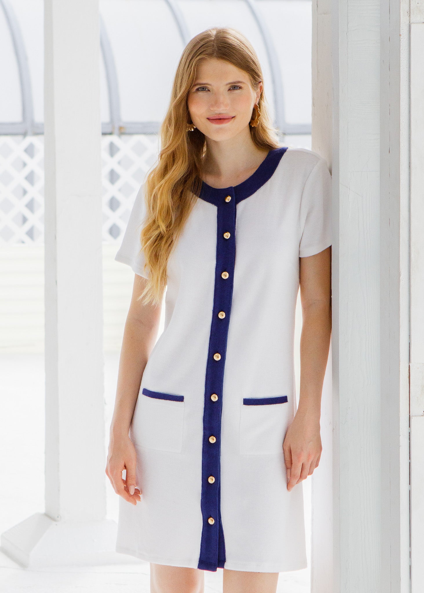 Woman wearing a white and blue terry dress with gold buttons standing outside a white cottage.