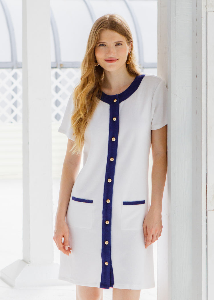Woman wearing a white and blue terry dress with gold buttons standing outside a white cottage.