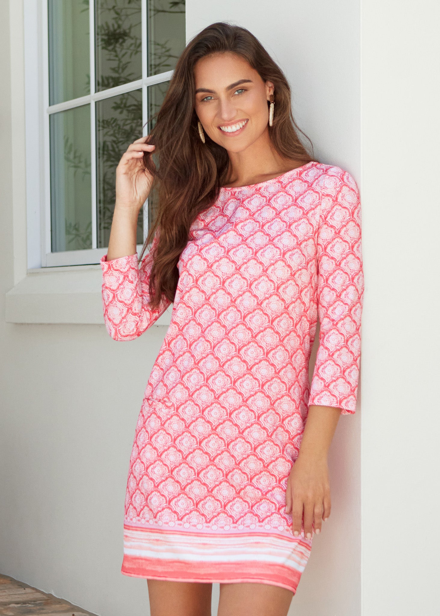 Woman wearing a pink patterned dress standing against a white wall.