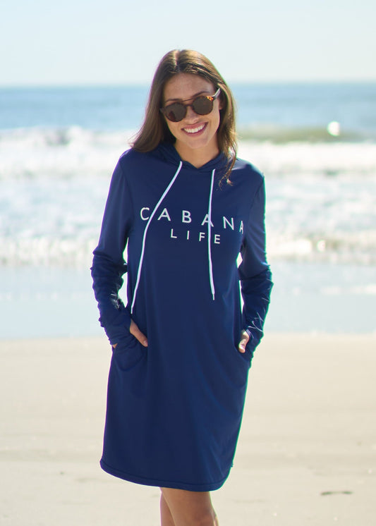 A woman smiling on the beach wearing the Navy Cabana Life Hoodie Dress with hands in pockets.