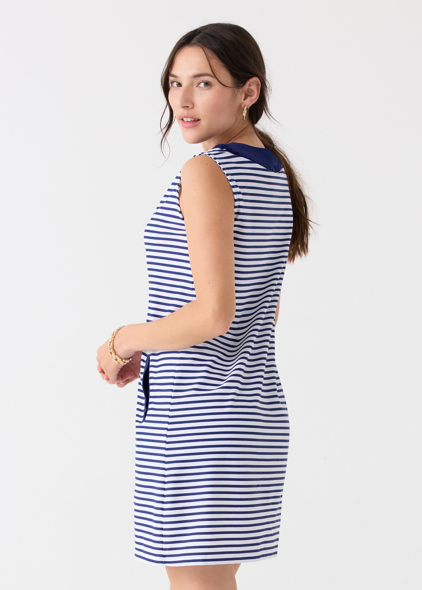 A dark haired woman looking back while wearing the Cabana Life sun protective Navy Stripe Sleeveless Fringe Tunic Dress with gold hoop earrings.