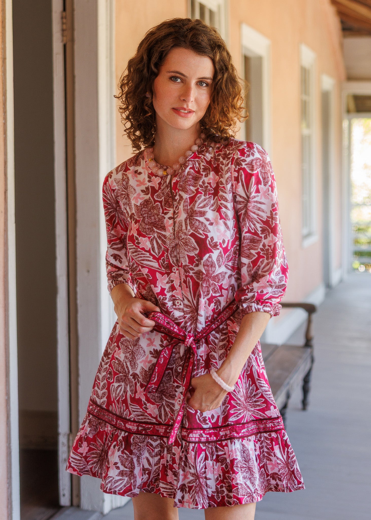 A woman with curly brown hair stands on a porch wearing a pink and white floral print mini dress with a tie waist and ruffled hem.