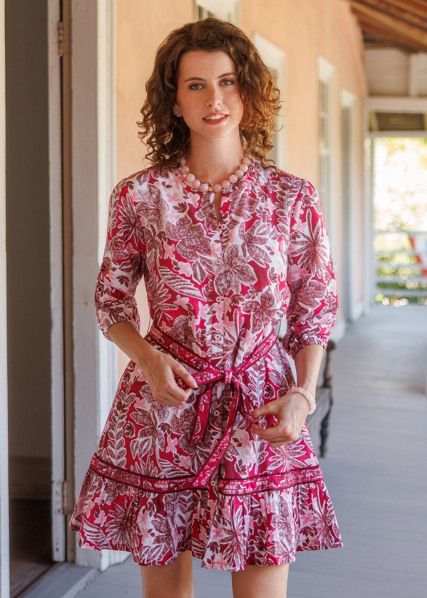 A woman wearing a pink and white floral print mini dress with a tie waist, standing on a covered porch.