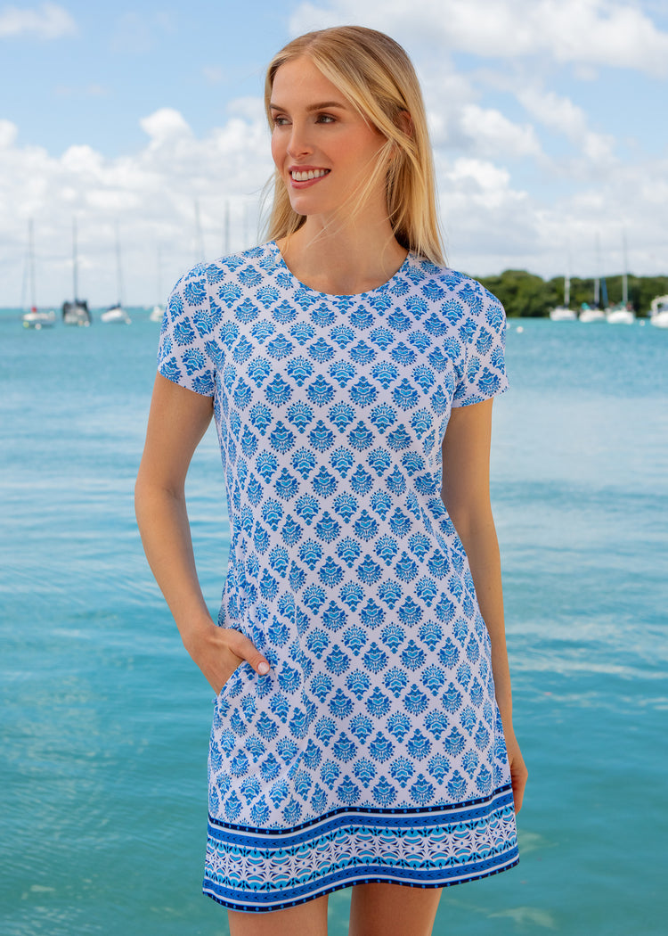 Woman wearing a blue and white patterned dress standing by a body of water with boats in the background.