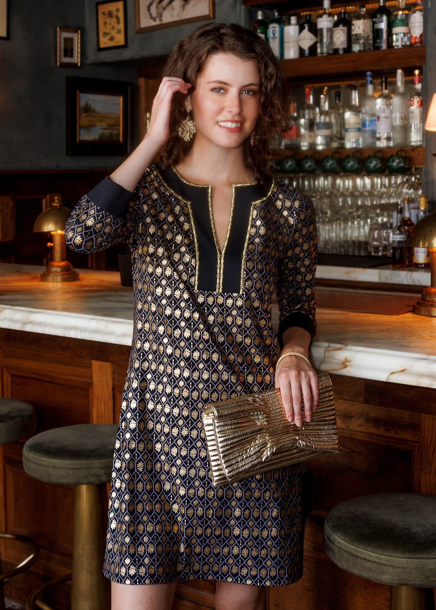 A woman stands in a bar setting wearing a navy and gold patterned tunic dress with a geometric print, holding a metallic gold clutch.