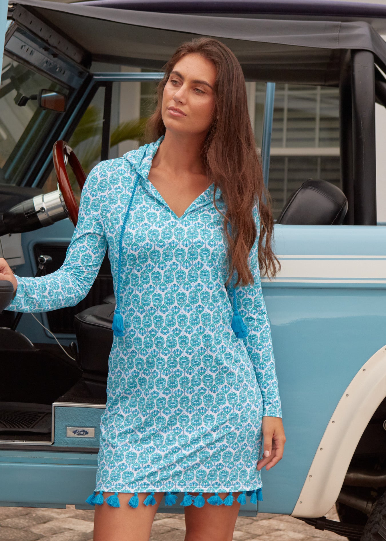 Woman wearing a blue patterned dress with tassel trim standing next to a vintage car.