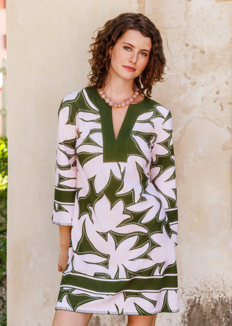 A woman wearing a short, three-quarter sleeve tunic dress with a bold green and pink floral pattern, standing against a textured beige wall.