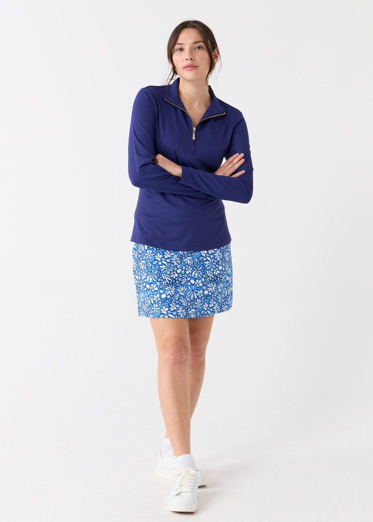 A dark haired woman with her arms crossed wearing the Cabana Life sun protective Navy 1/4 Zip Sport Top and Seaside Floral 17" Skort with white crew socks and sneakers.