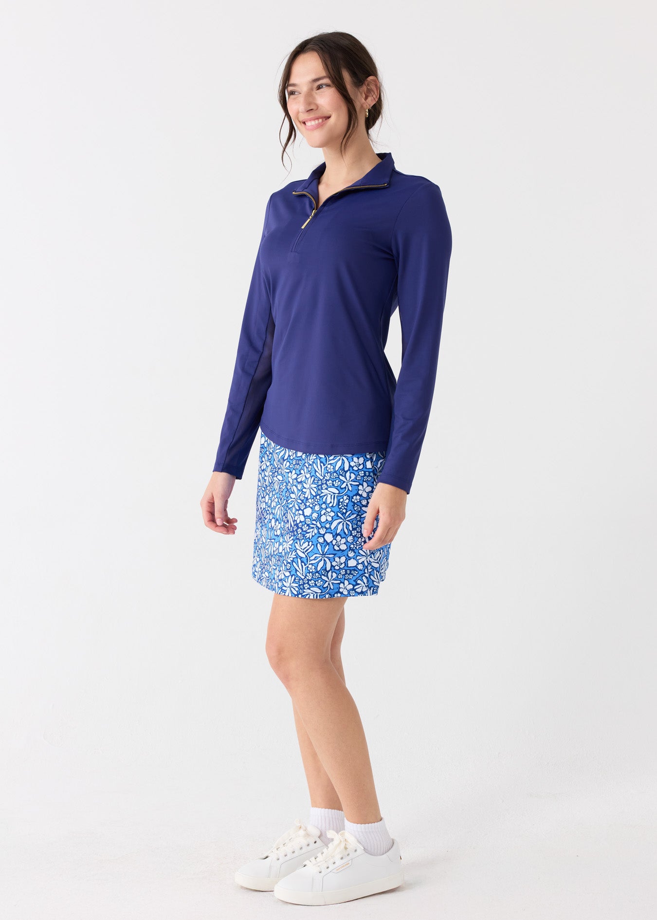 The side of a dark haired woman wearing the Cabana Life sun protective Navy 1/4 Zip Sport Top and Seaside Floral 17" Skort with white crew socks and sneakers.