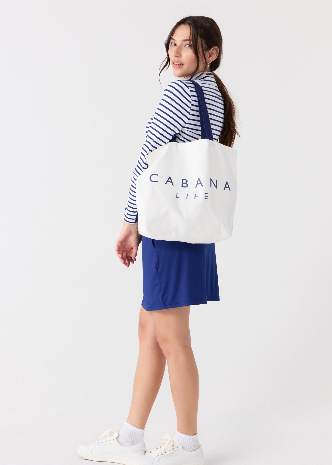 A woman looking back with the Cabana Like Tote Bag on her shoulder and wearing the sun protective Navy Stripe 1/4 Zip Sport Top with the Navy 17" Skort.