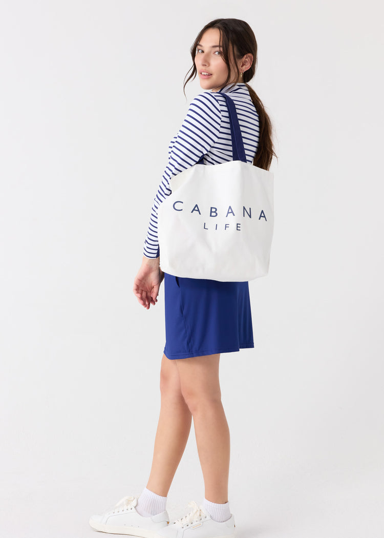 A woman looking back with the Cabana Like Tote Bag on her shoulder and wearing the sun protective Navy Stripe 1/4 Zip Sport Top with the Navy 17" Skort.