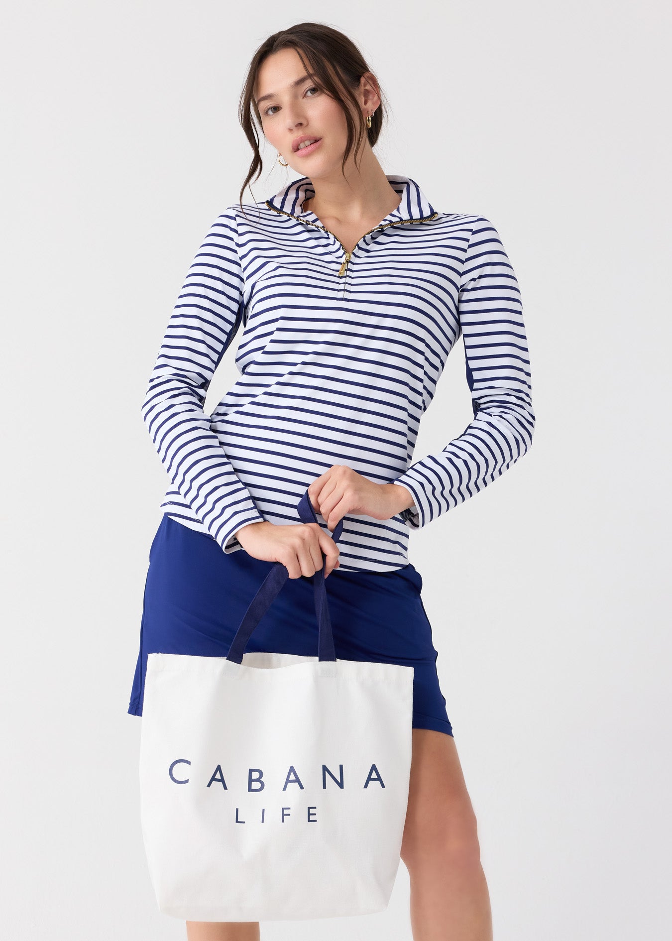 A woman wearing the sun protective Navy Stripe 1/4 Zip Sport Top and Navy 17" Skort while holding the Cabana Life Tote Bag.
