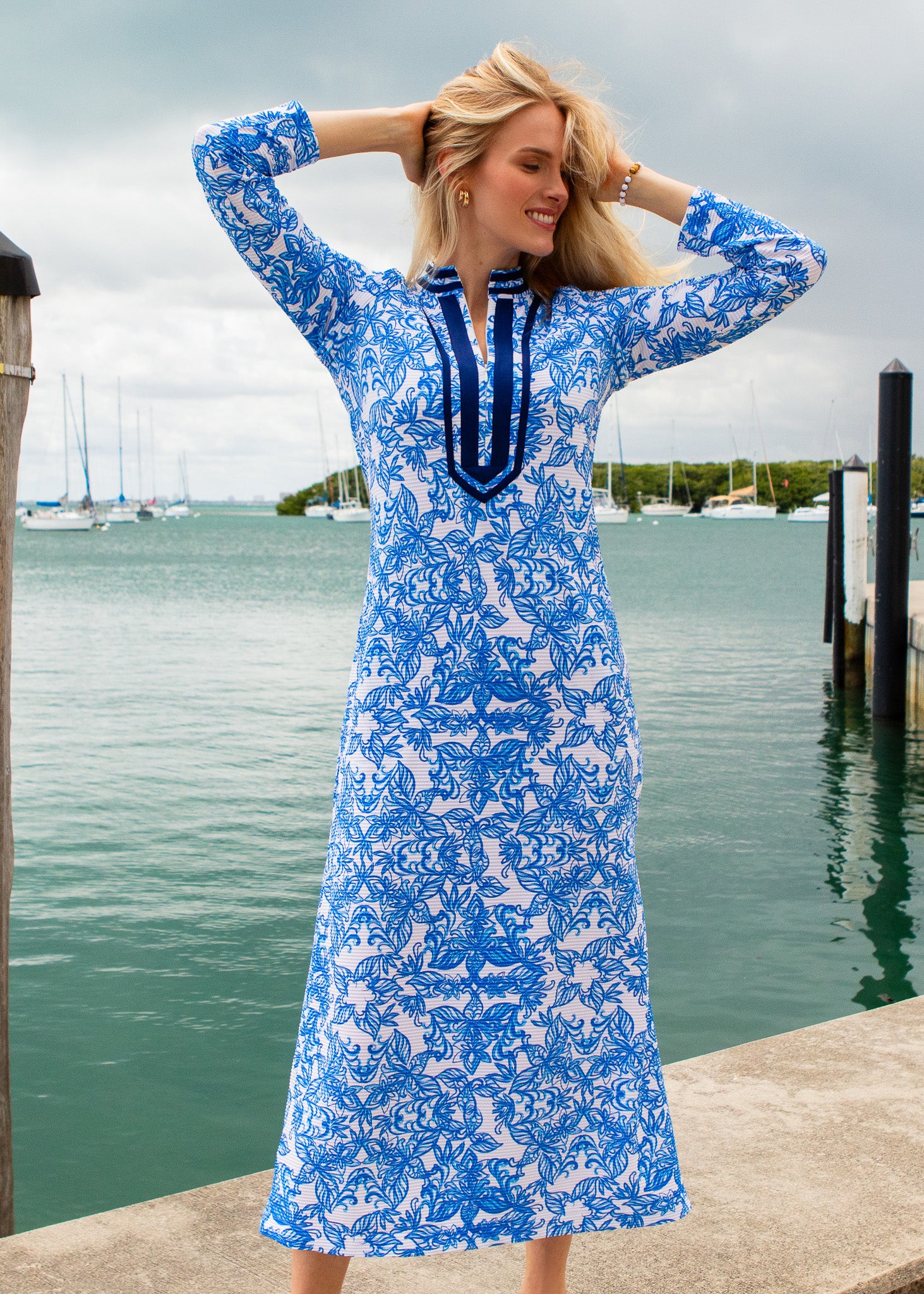 Woman in a blue floral dress standing by a dock with water and boats in the background