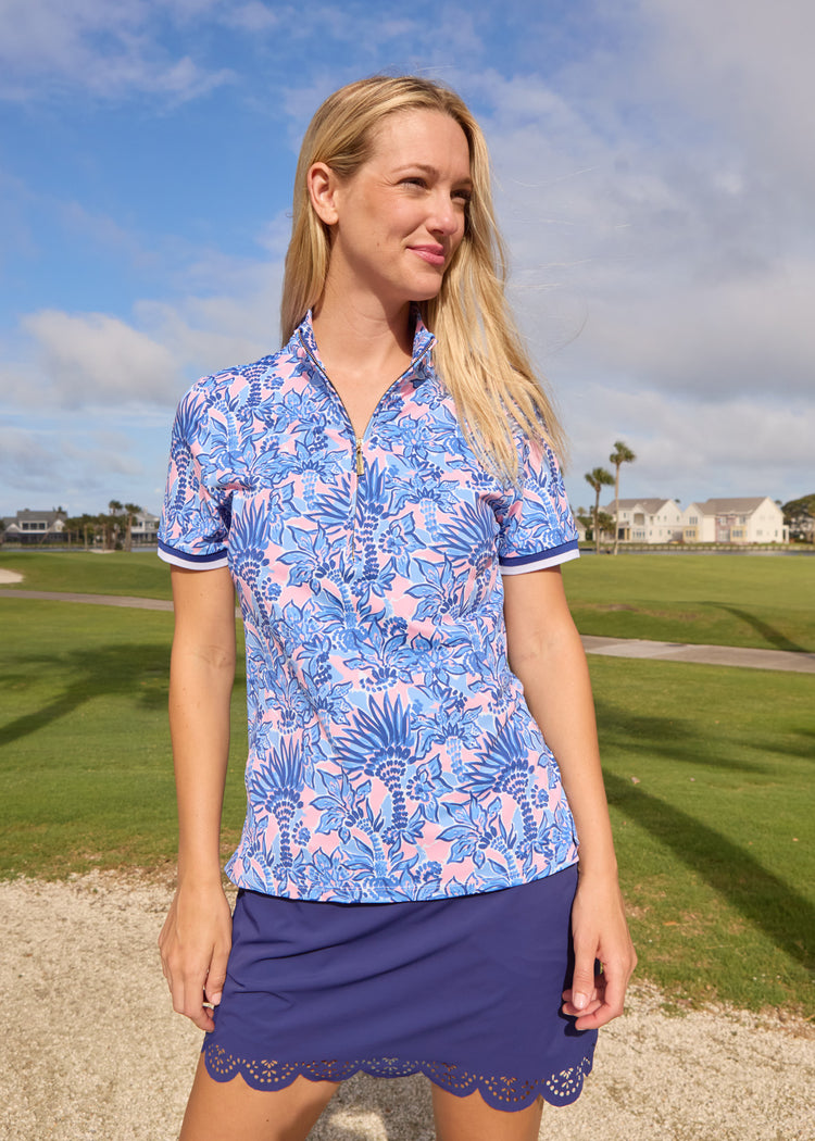 A blonde woman wearing the Cabana Life sun protective Palm Beach Short Sleeve Collared 1/4 Zip with the Navy 15" Scallop Skort in front of a golf course.