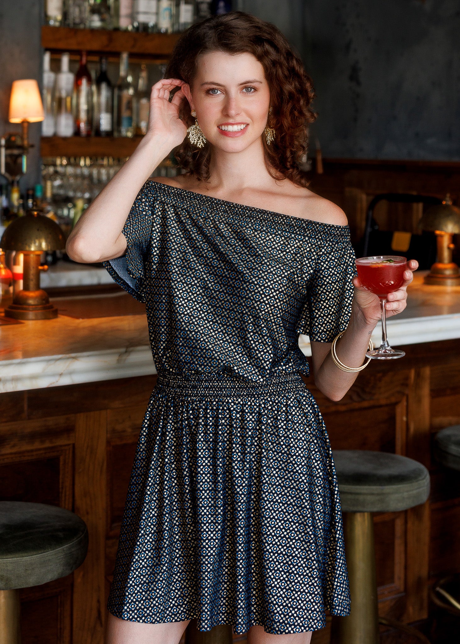 A woman with curly brown hair stands in a bar setting wearing a black metallic print off-the-shoulder dress. She is leaning on a marble bar counter with dark wood stools in front of a liquor shelf.