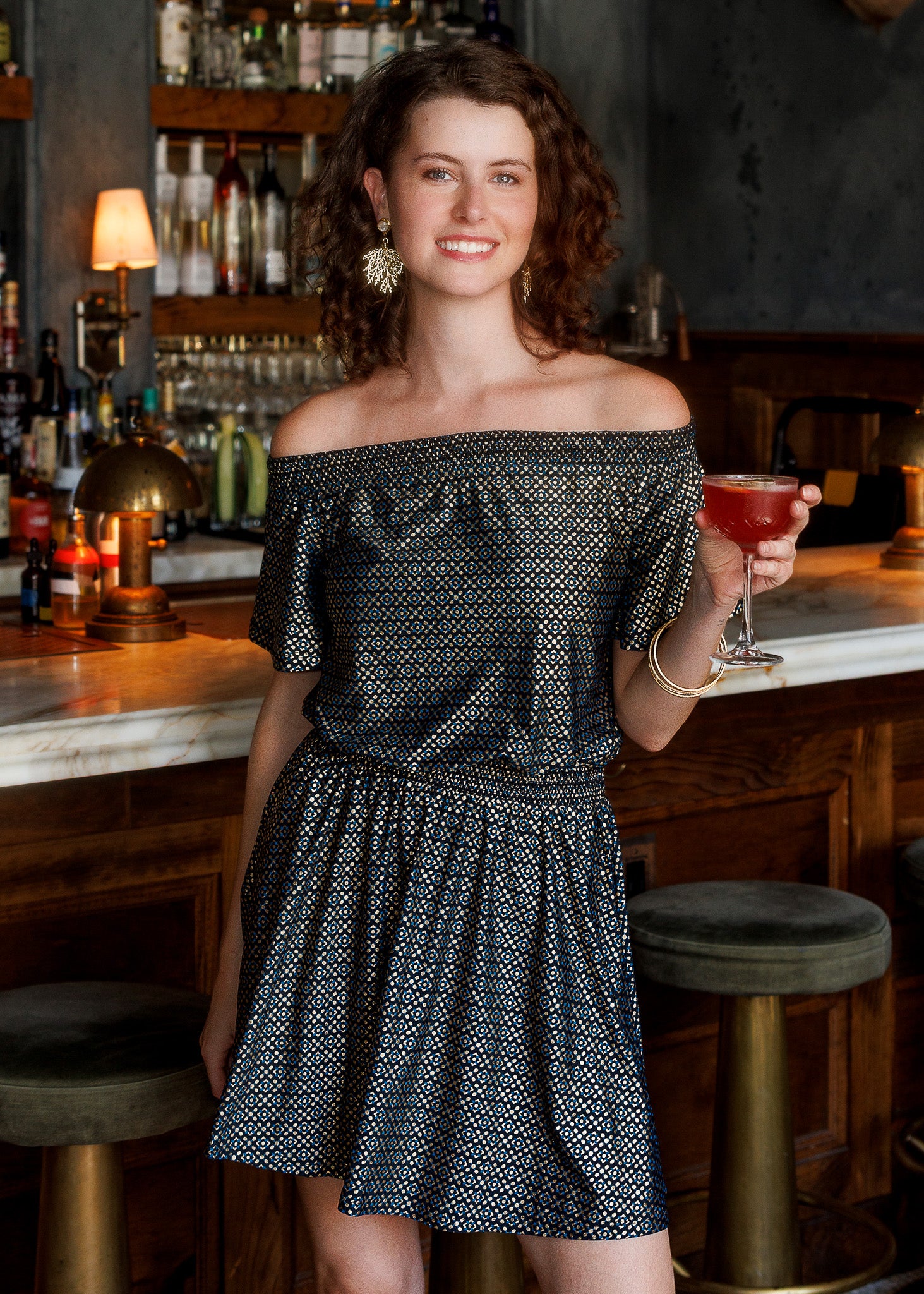 A woman with curly brown hair stands in a bar setting holding a pink cocktail wearing a black metallic print off-the-shoulder dress. She is leaning on a marble bar counter with dark wood stools in front of a liquor shelf.