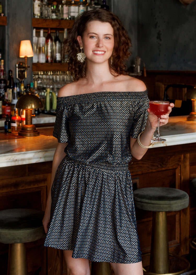 A woman with curly brown hair stands in a bar setting holding a pink cocktail wearing a black metallic print off-the-shoulder dress. She is leaning on a marble bar counter with dark wood stools in front of a liquor shelf.