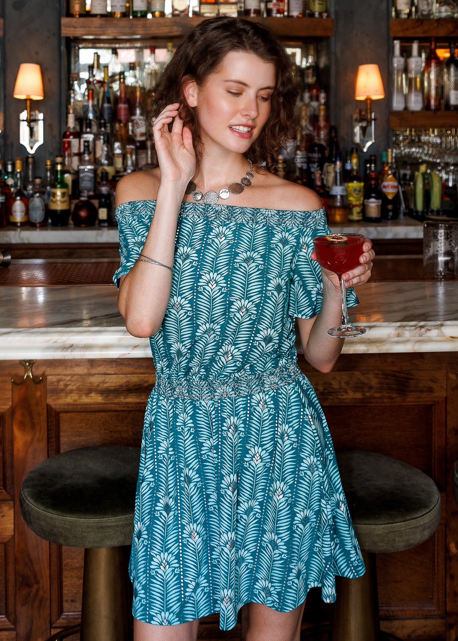 A woman wearing a teal and white palm leaf print off-the-shoulder dress stands in a bar setting, holding a red cocktail glass.