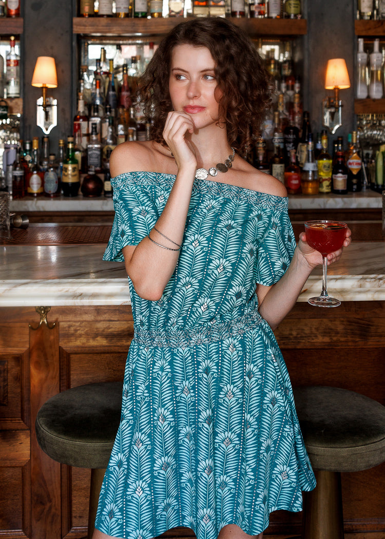 A woman wearing a teal and white palm leaf print off-the-shoulder dress stands in a bar setting, holding a red cocktail glass.