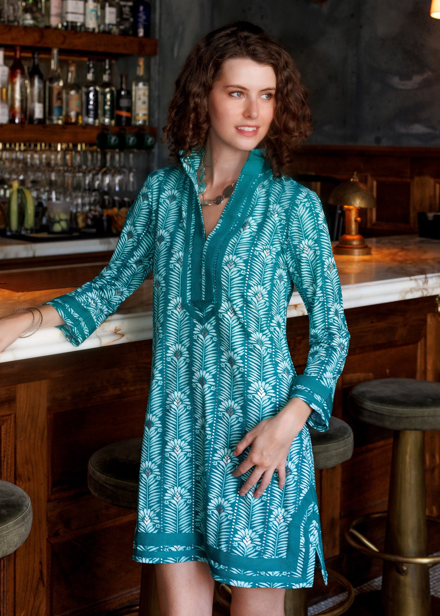 A woman wearing a teal and white printed dress stands in a bar setting with wooden stools and shelves of bottles in the background.