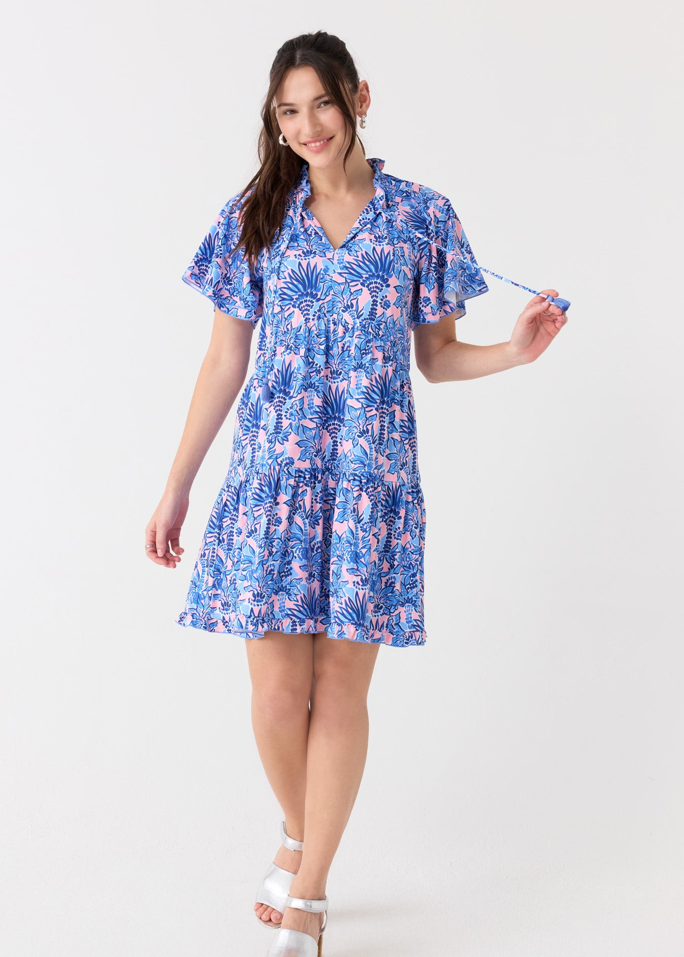 A brunette woman holding the tassel tie on the Cabana Life sun protective Palm Beach Short Sleeve Tiered Dress, paired with silver sandals.