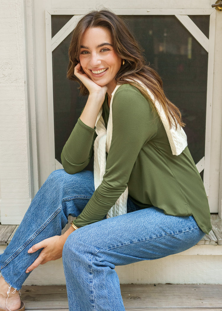 Woman sitting on a wooden step wearing a green sweater and blue jeans, smiling.