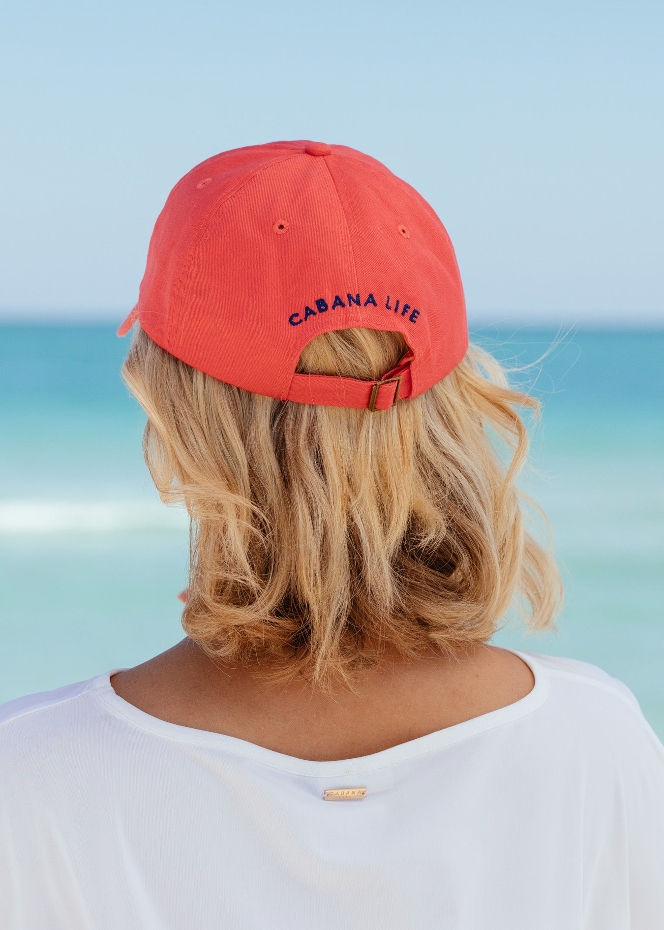 Back of blonde woman wearing Nantucket Red Cabana Life Baseball Hat with white rashguard.