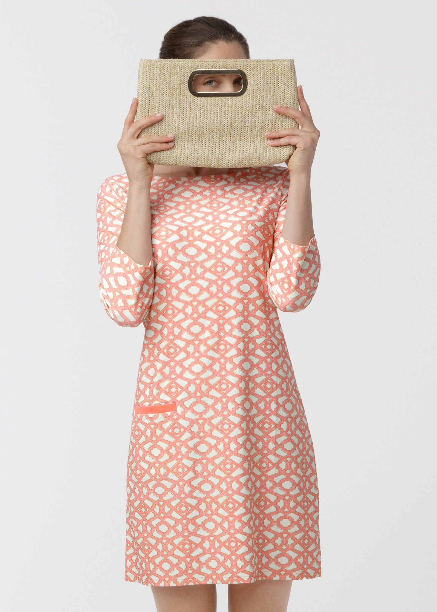 Woman wearing a pink patterned dress holding a woven bag over her face on a white background