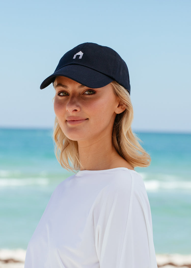 Blonde woman wearing Navy Cabana Life Baseball Hat with white rashguard on beach.