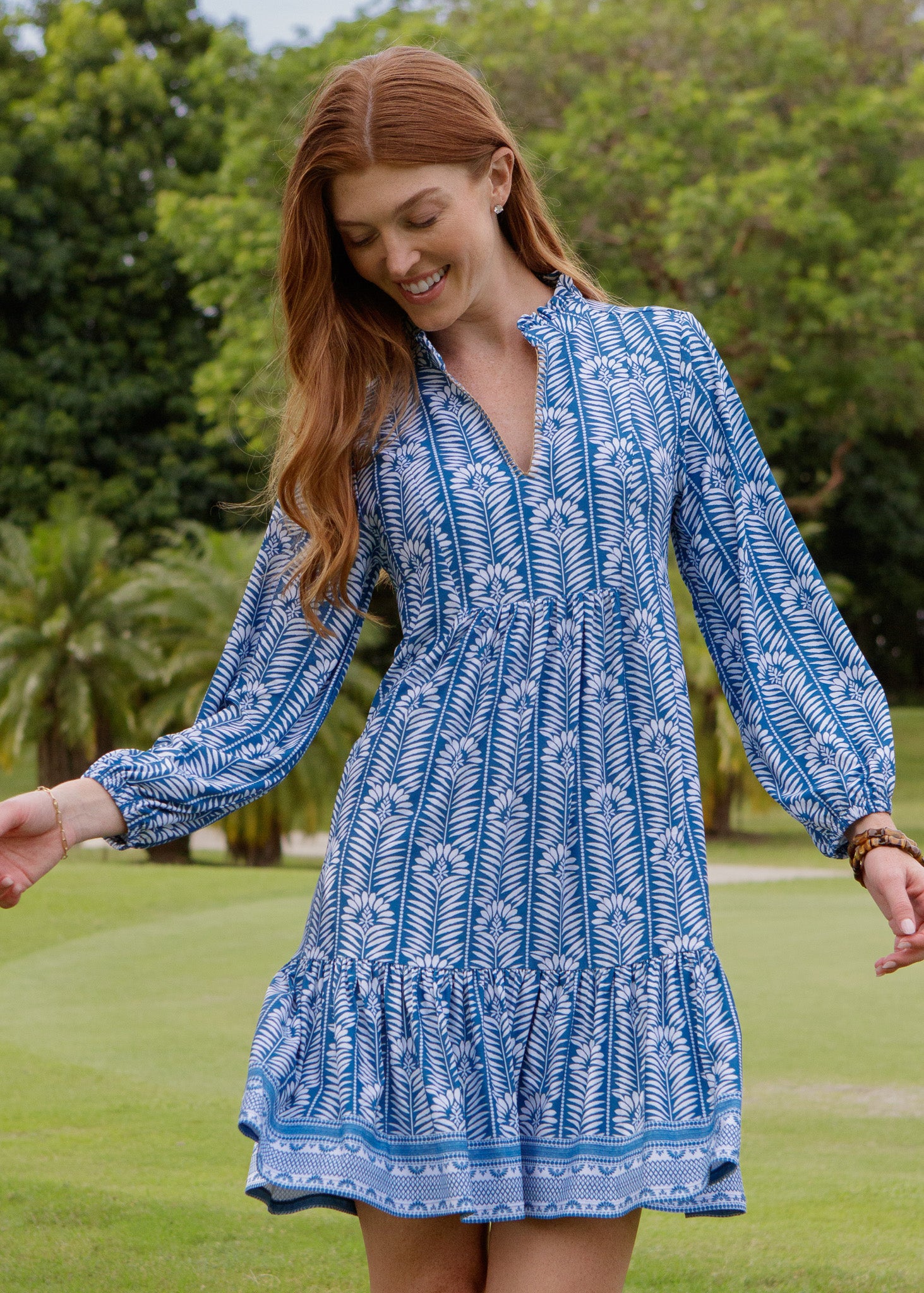 Woman wearing a blue patterned dress standing in a grassy area with trees in the background