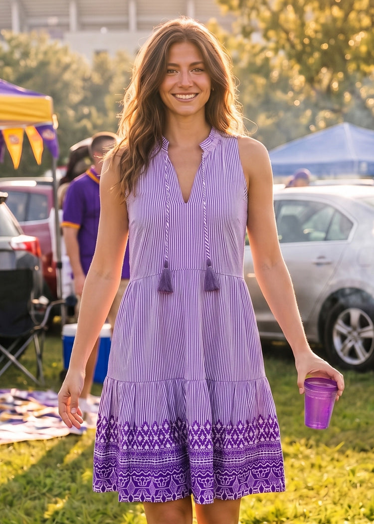 A woman wearing a sleeveless purple and white vertically striped tiered dress with tassel ties and a purple geometric patterned hem, standing outdoors at a sunny event and holding a purple cup.