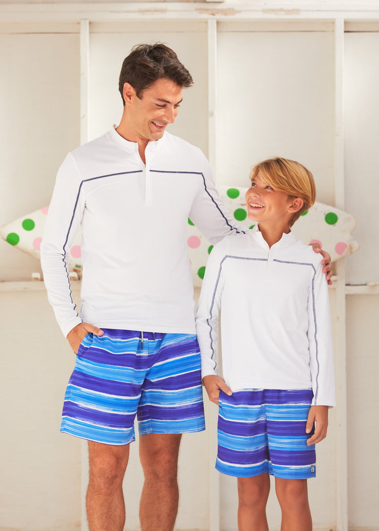 Man and boy both standing in front of a polka dotted surfboard. The man is wearing the Cabana Life sun protective San Sebastian Stripe Men's Swim Trunk. 